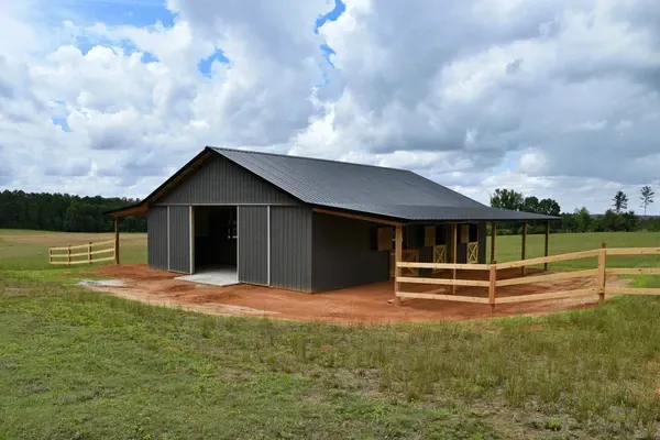 New gray barn-style house with wooden porch and fence in a grassy field under cloudy sky