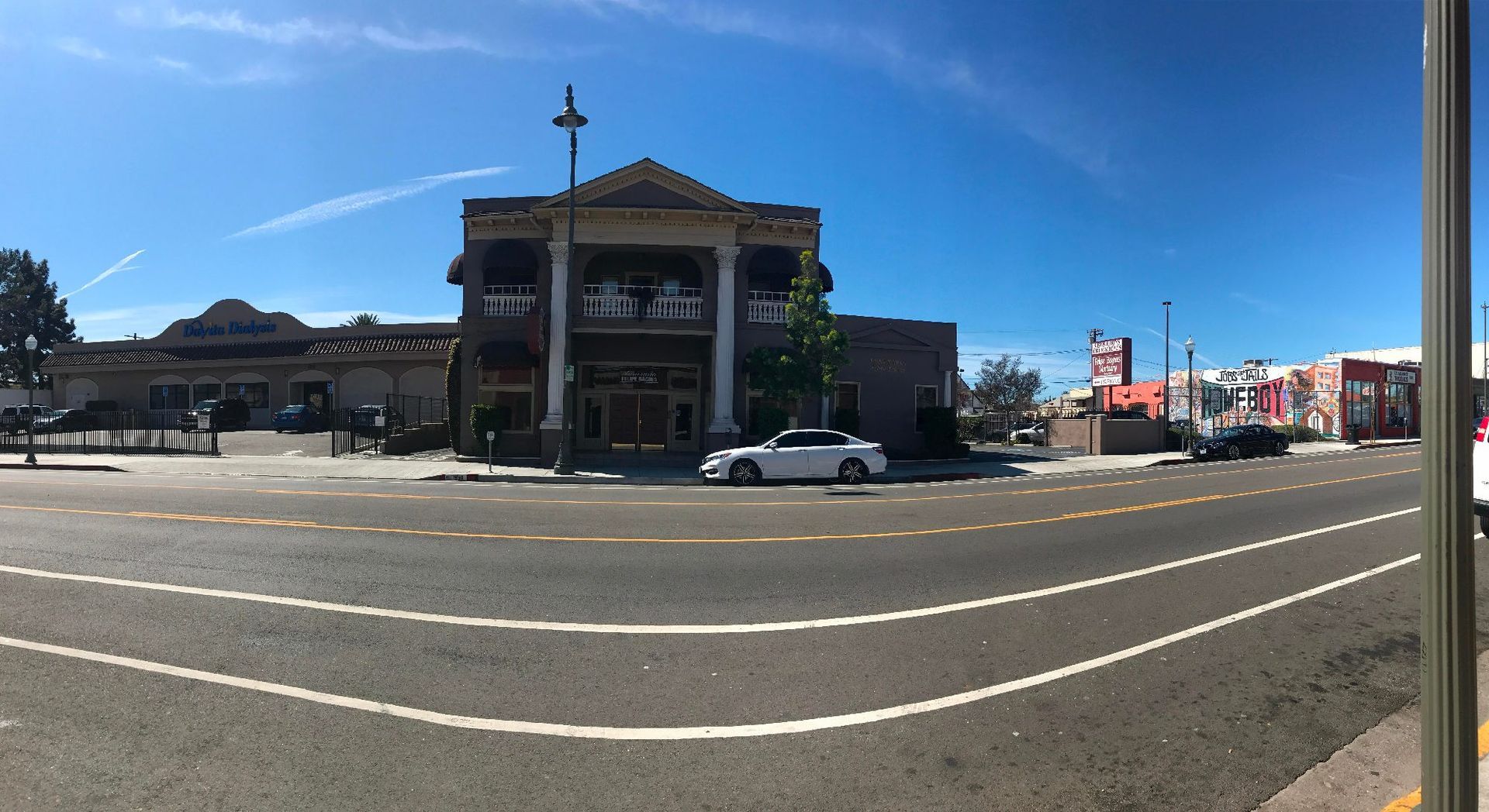 A street-level panorama showing a multi-story historic building with columns under a bright blue sky on a sunny day.