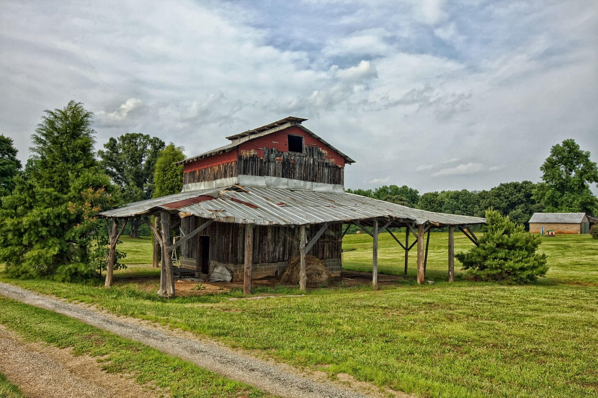Old weathered barn with red roof in a green field under cloudy sky.