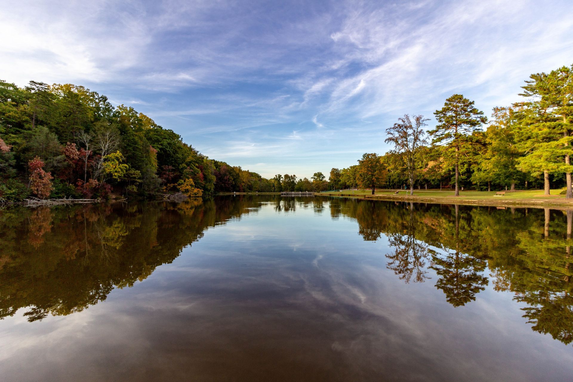 Lake scene with trees lining the banks, reflecting in the calm water under a cloudy blue sky.
