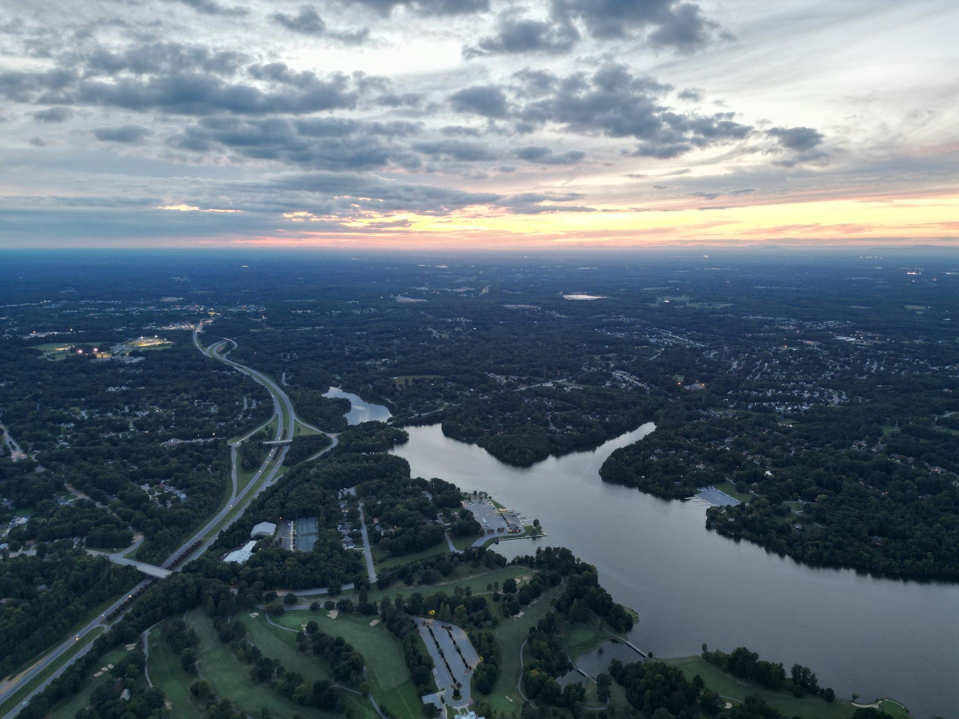 Aerial view of a lake and green trees with a highway and a cloudy sky at sunset.