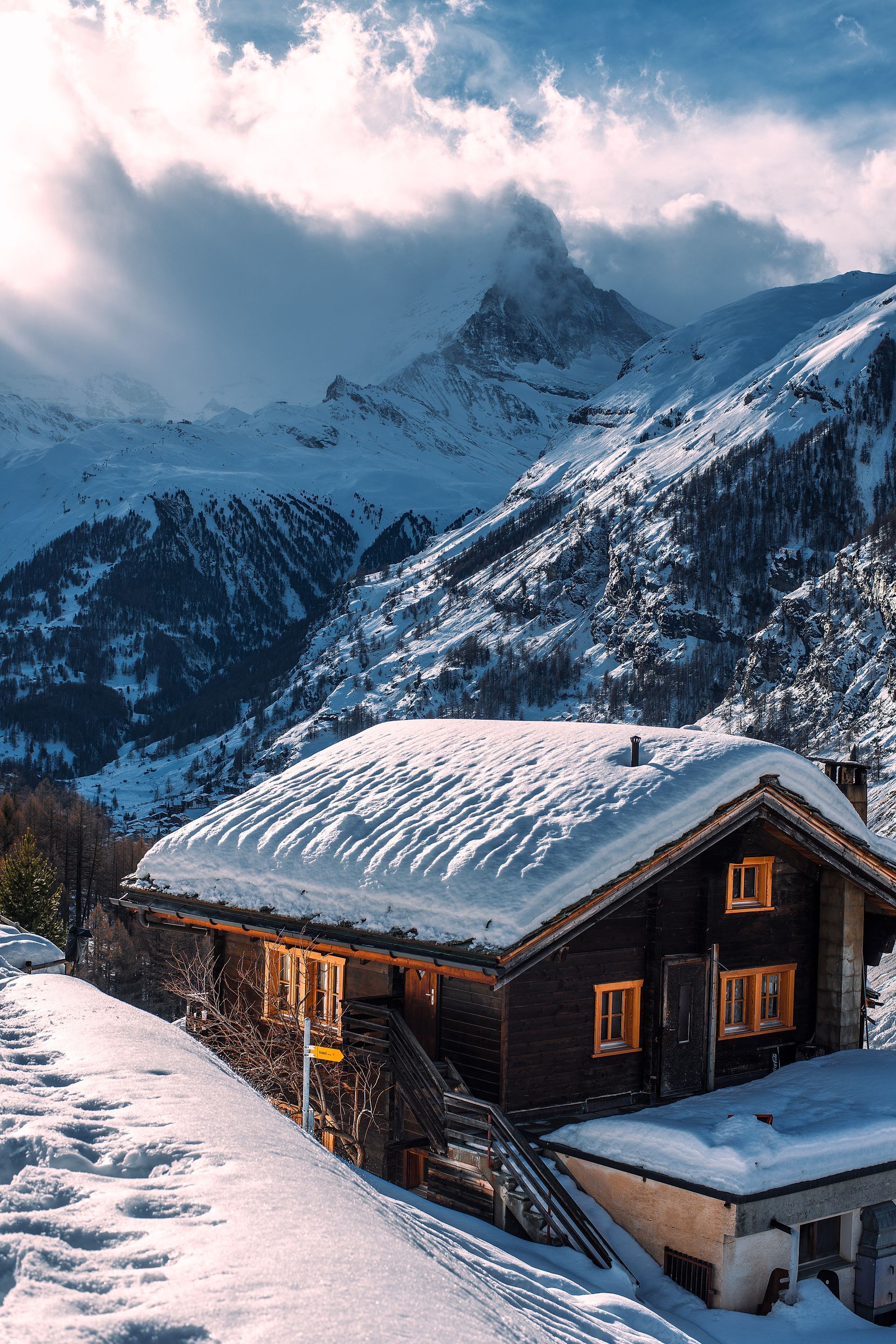 Snow-covered chalet nestled at the base of snowy mountains, with a bright sky breaking through the clouds.