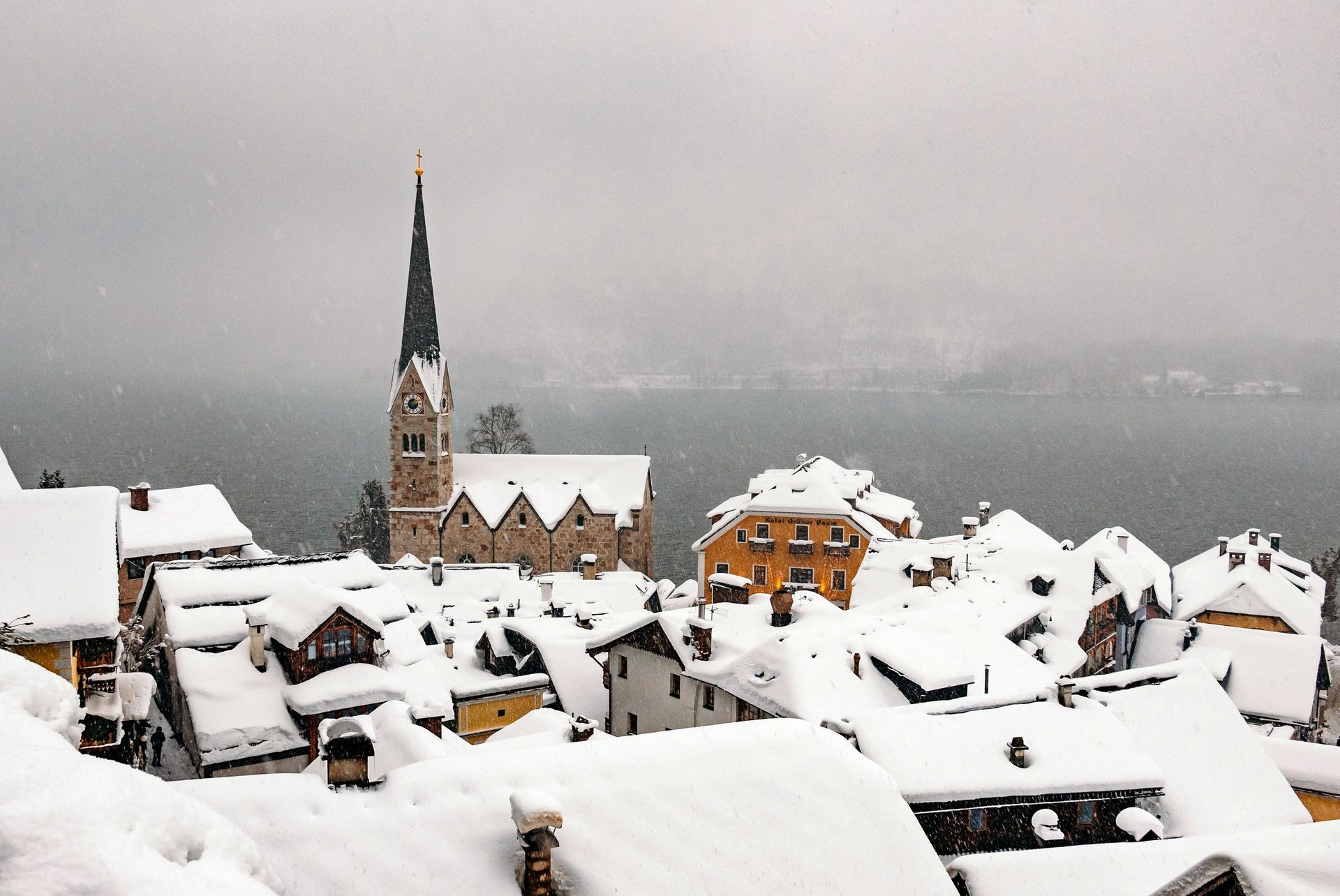 Snow-covered rooftops and a church spire in a village by a lake under a gray sky.