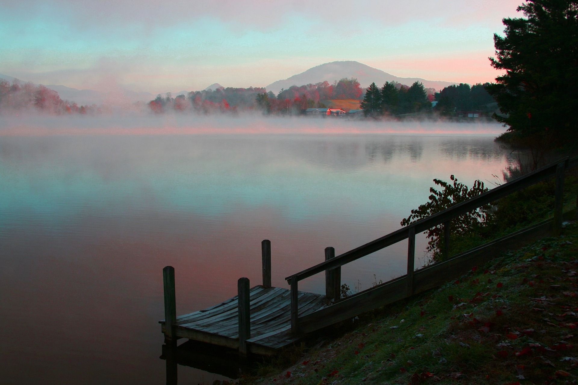 A tranquil lake scene at dusk. Fog drifts over the water, with a small dock in the foreground. Pink and blue hues.