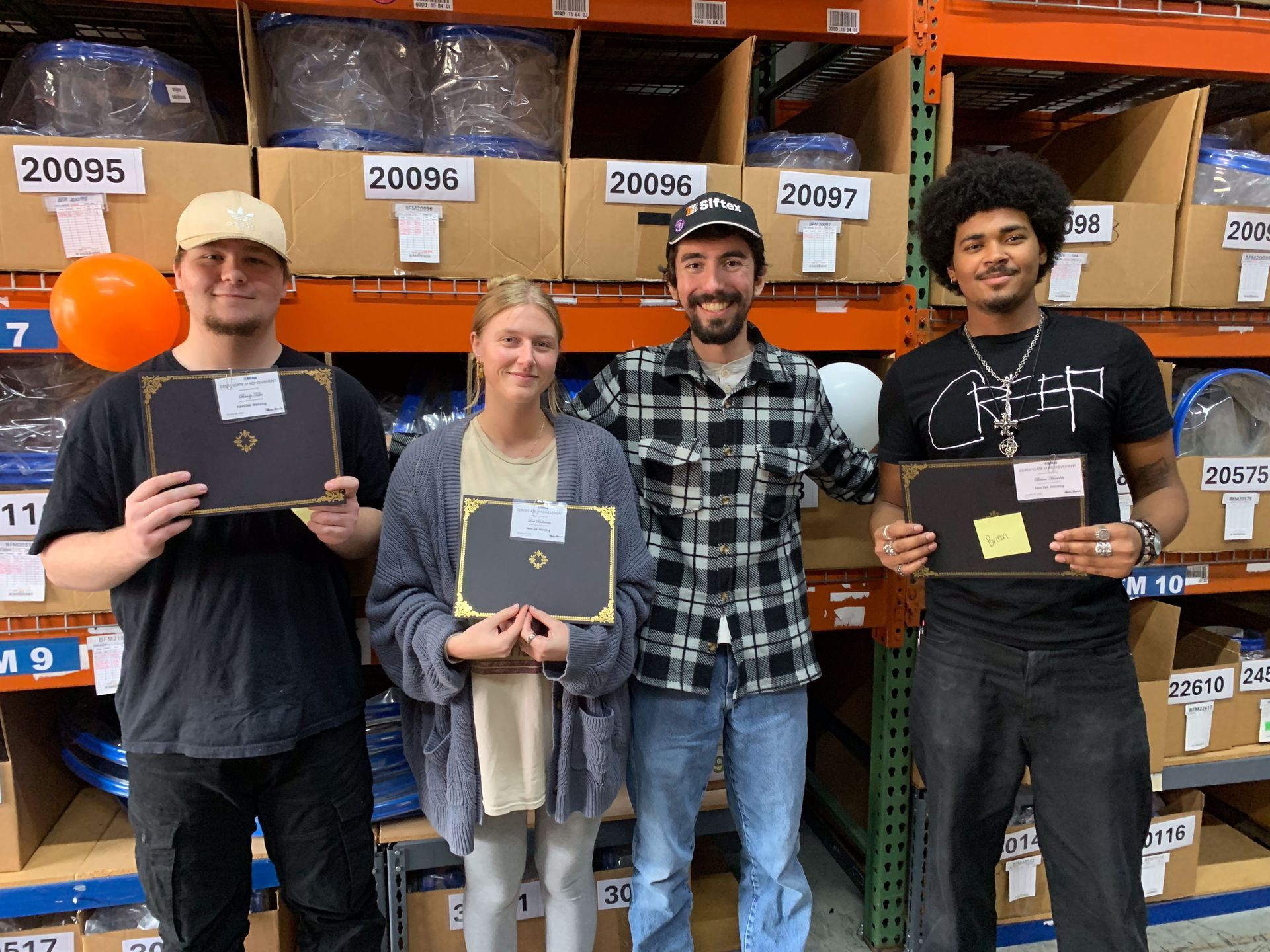 Four people holding plaques in a warehouse aisle with labeled boxes on shelves behind them