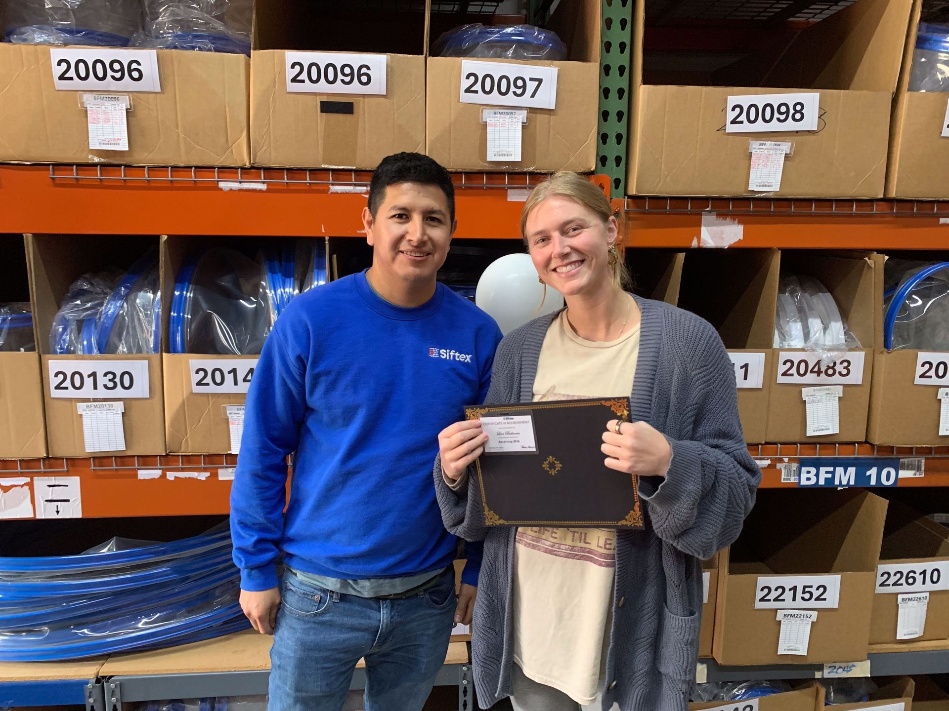 Two people stand in a warehouse aisle holding a framed item, with shelves of labeled boxes behind them.