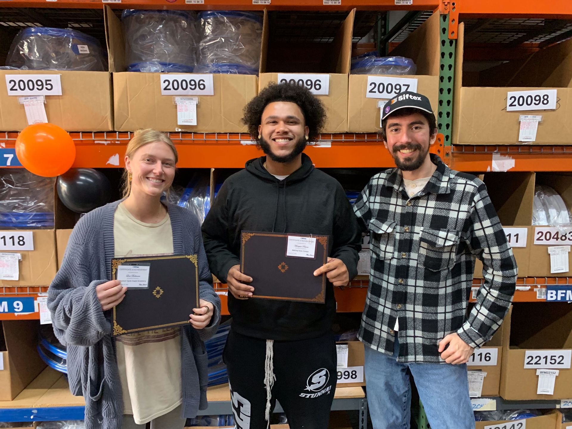 Three people smiling in a warehouse aisle, holding plaques in front of orange shelving.