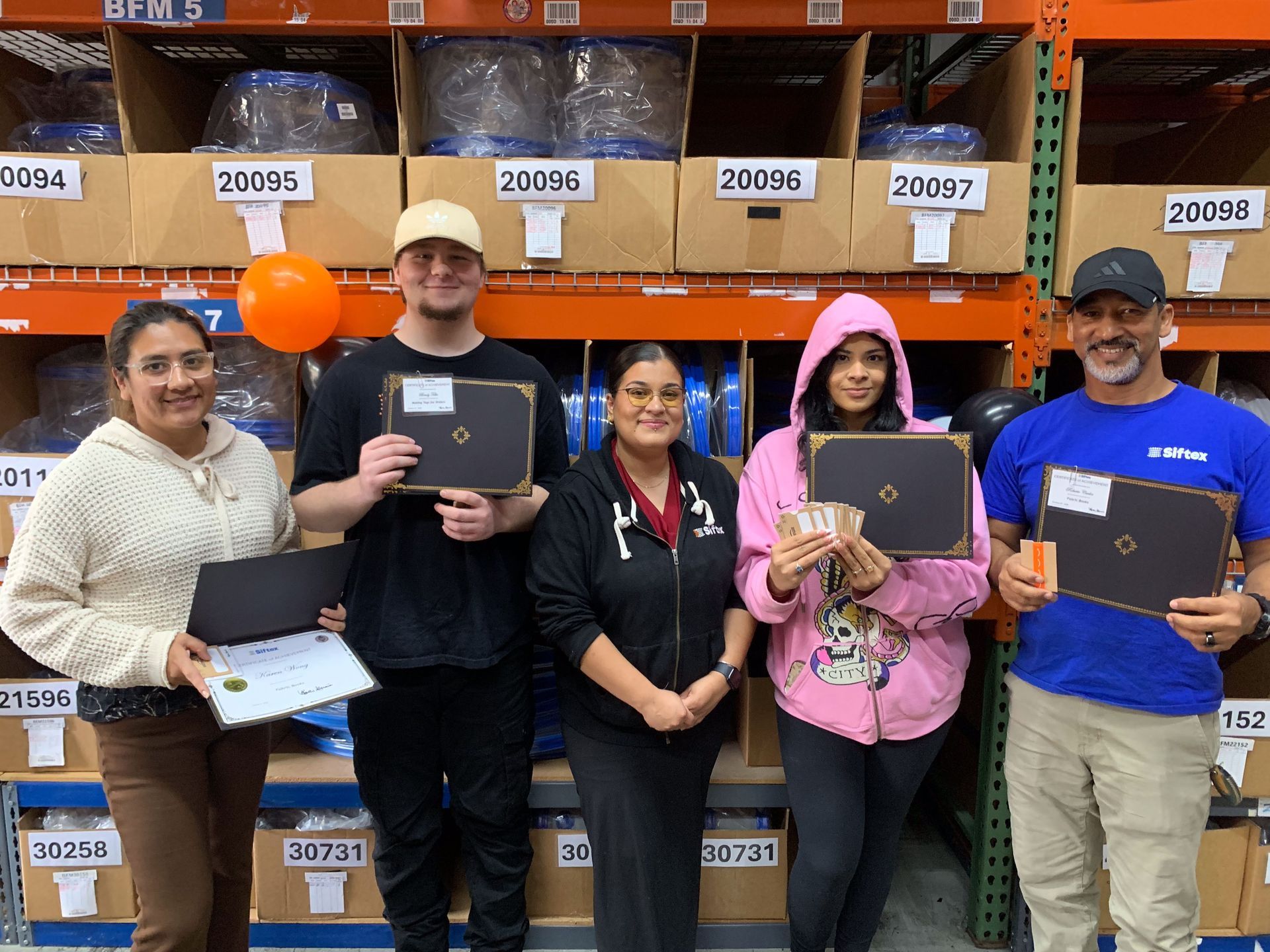 Five people pose in a warehouse aisle holding laptops and a plaque in front of shelves of labeled storage bins.