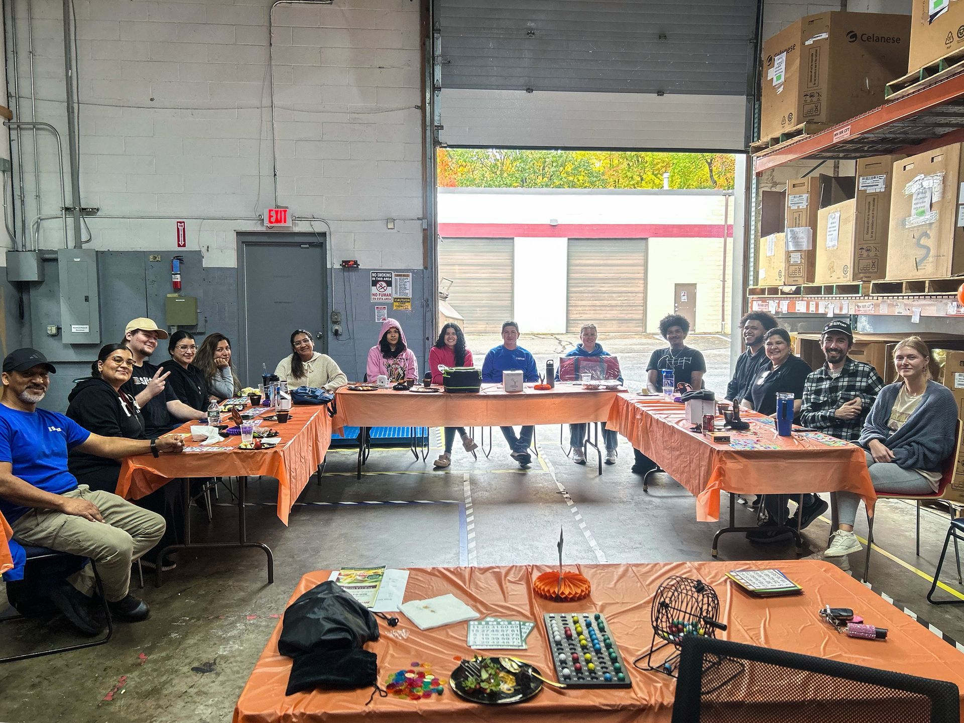 People seated around U-shaped tables in a warehouse meeting space, with orange tablecloths and supplies on tables.