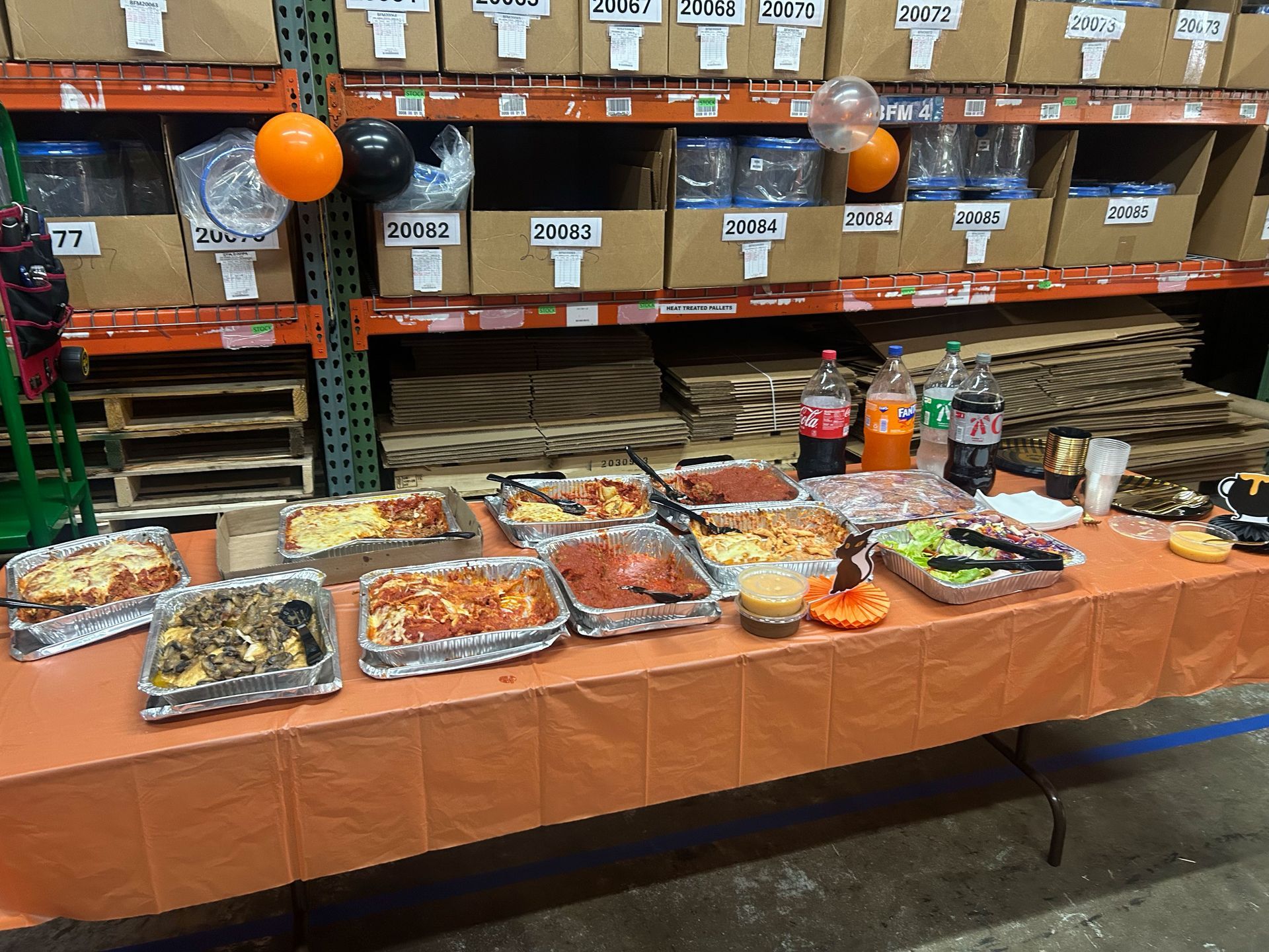 Buffet table with assorted dishes and drinks in a warehouse aisle, decorated with orange balloons and streamers