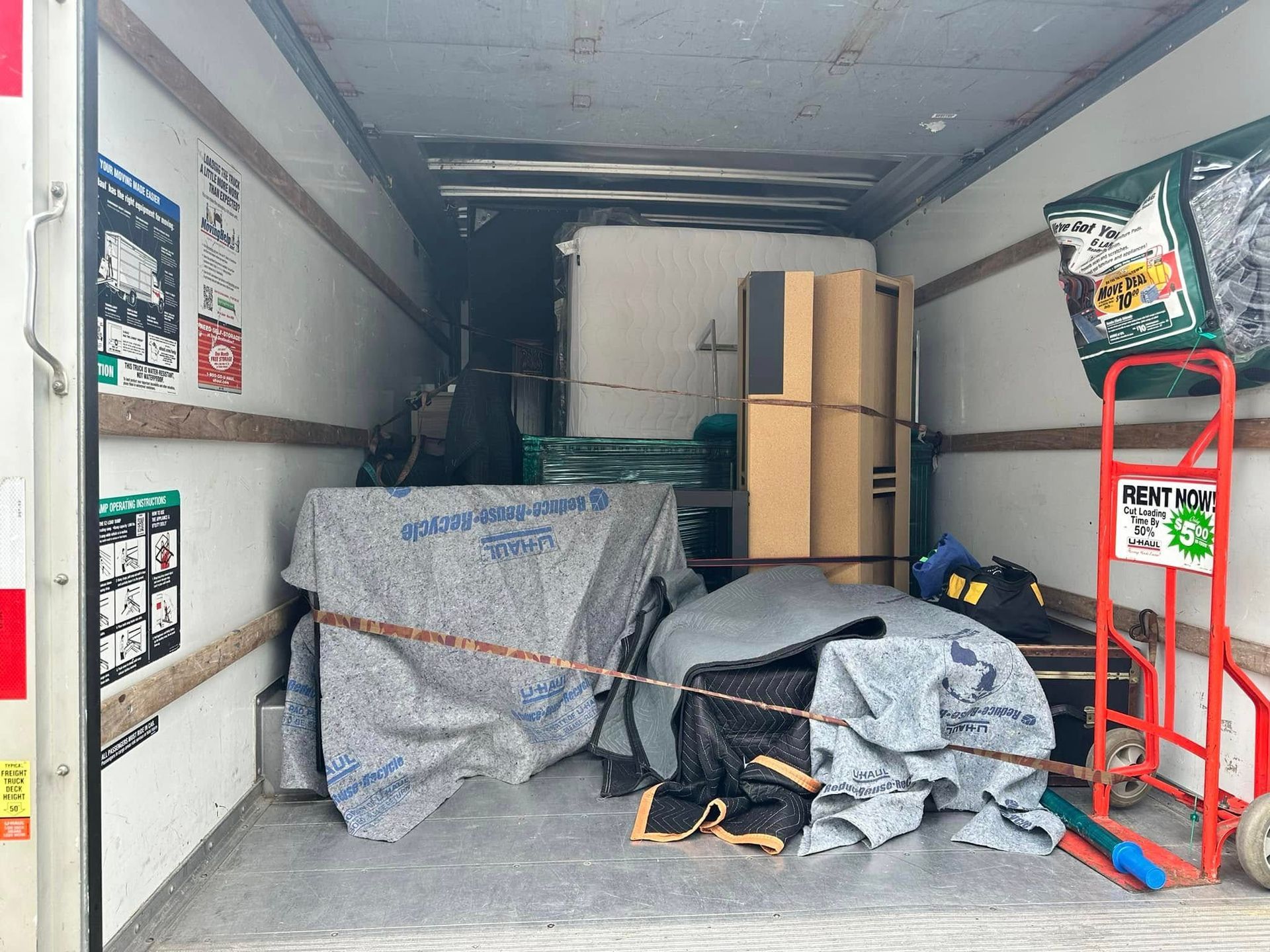 Interior of a moving truck partially filled with packed boxes, covered furniture, and a hand truck.