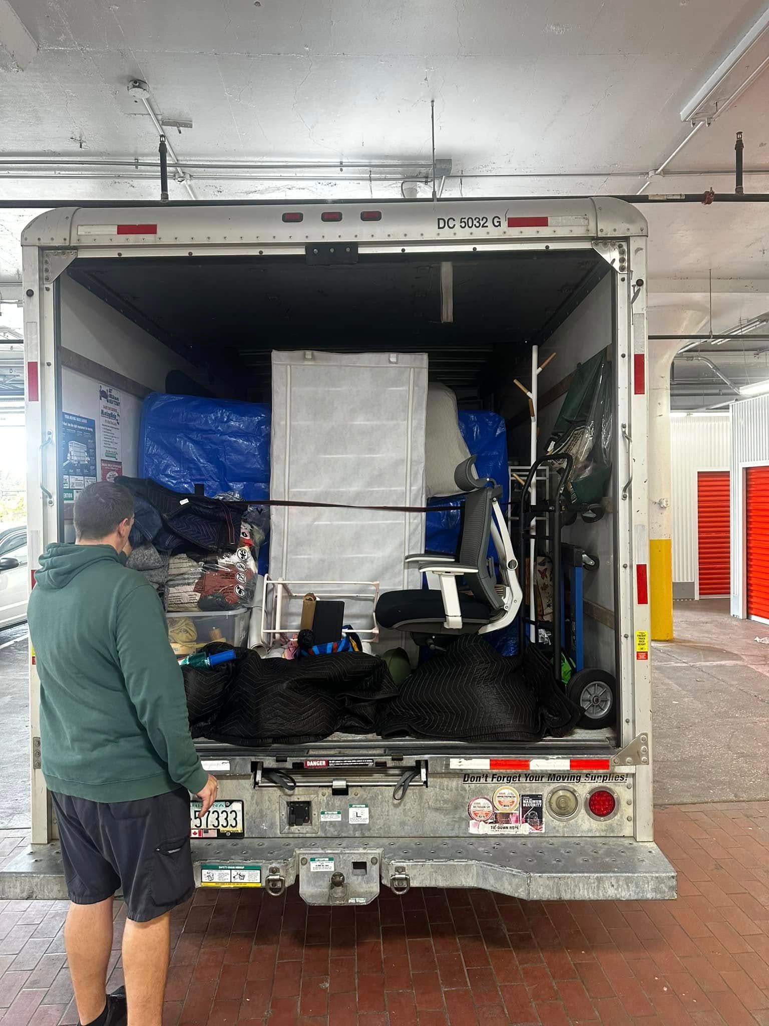 A person stands near the open back of a truck, loaded with various items and equipment.