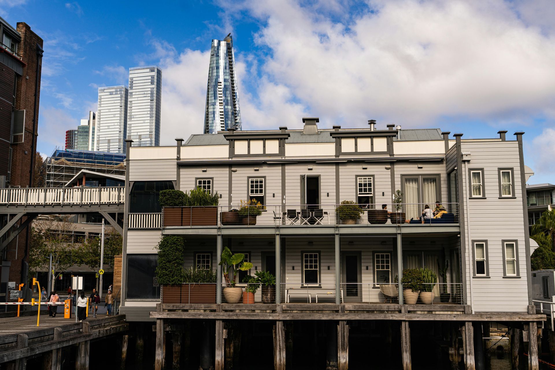 White waterfront building with balconies and potted plants, set against city skyscrapers and a partly cloudy sky