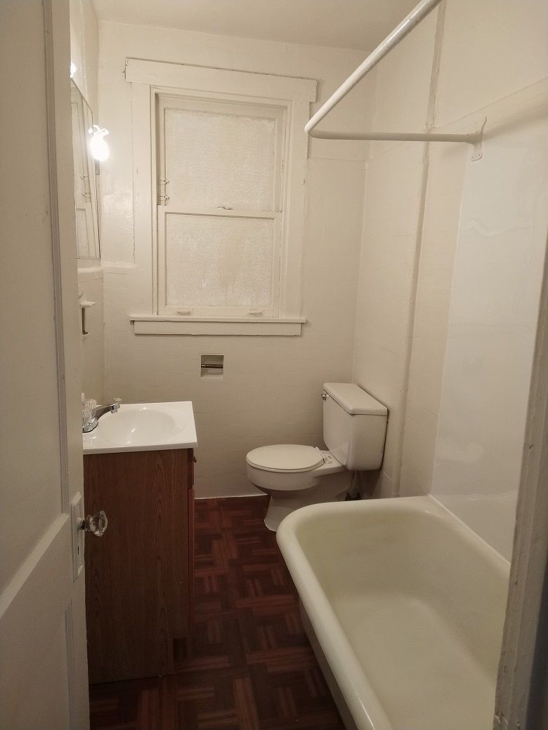 Small bathroom with a white tub, toilet, and sink. Brown wood-patterned floor and a window.