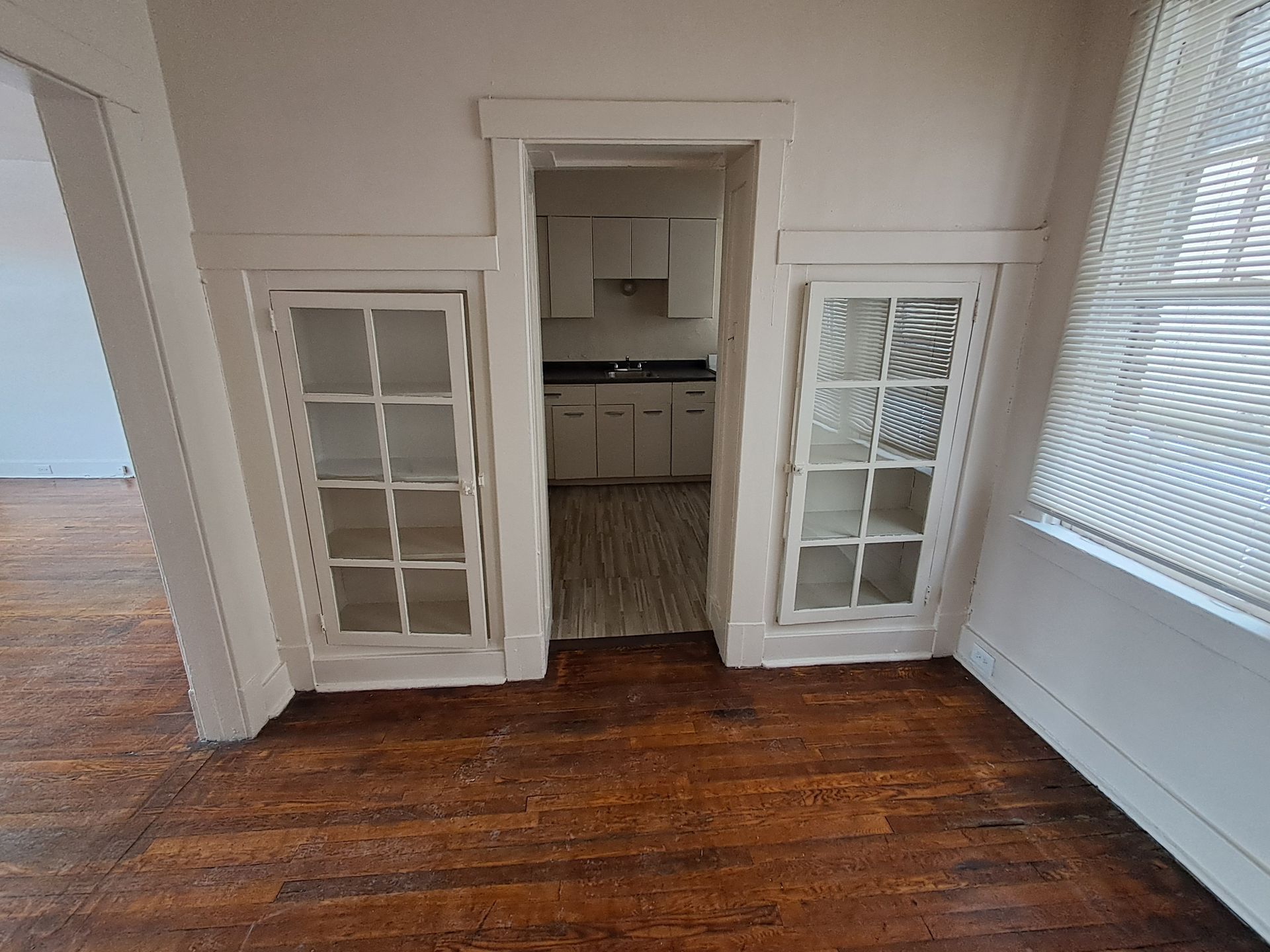 Interior with hardwood floors, built-in glass cabinets, and an opening to a kitchen with white cabinets.