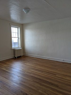Empty room with hardwood floors, radiator, and window. White walls, ceiling, and baseboards.