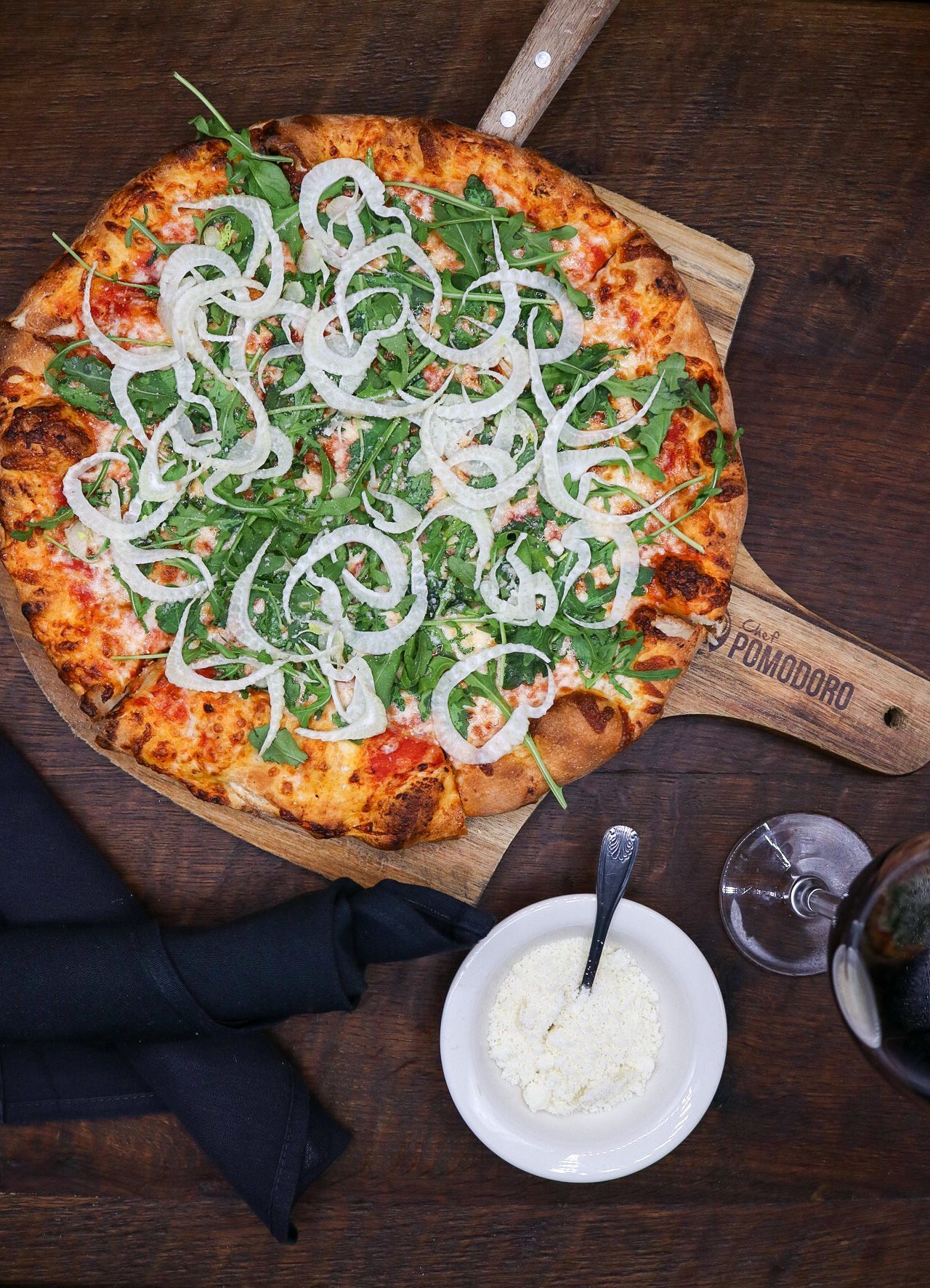 Pizza topped with onion rings and greens on a wooden board, with dipping sauce beside it.