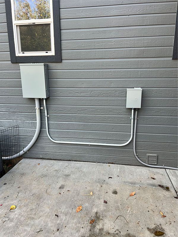 Gray house exterior with two utility boxes and conduit beneath a window above a concrete patio.