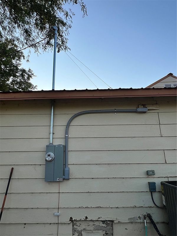 Exterior house wall with gray electrical meter box, conduit, and overhead wires under a blue sky