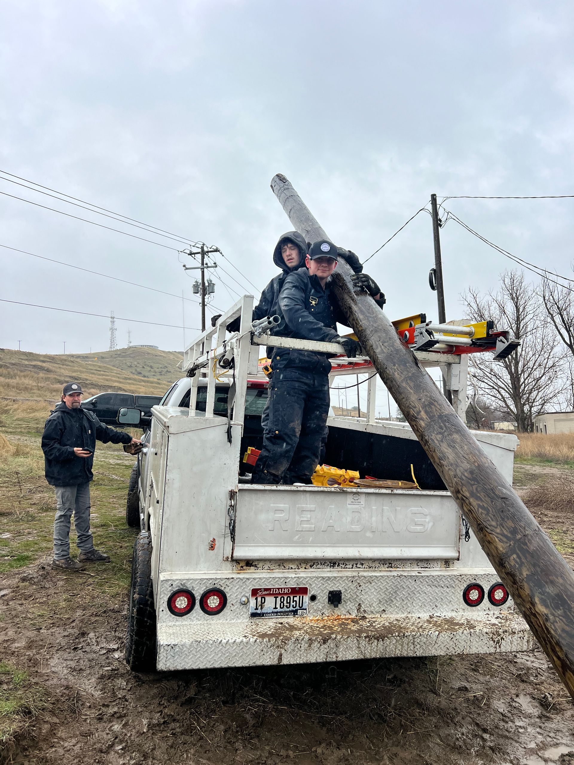 Workers load a utility truck with a long pole at a muddy roadside work site.