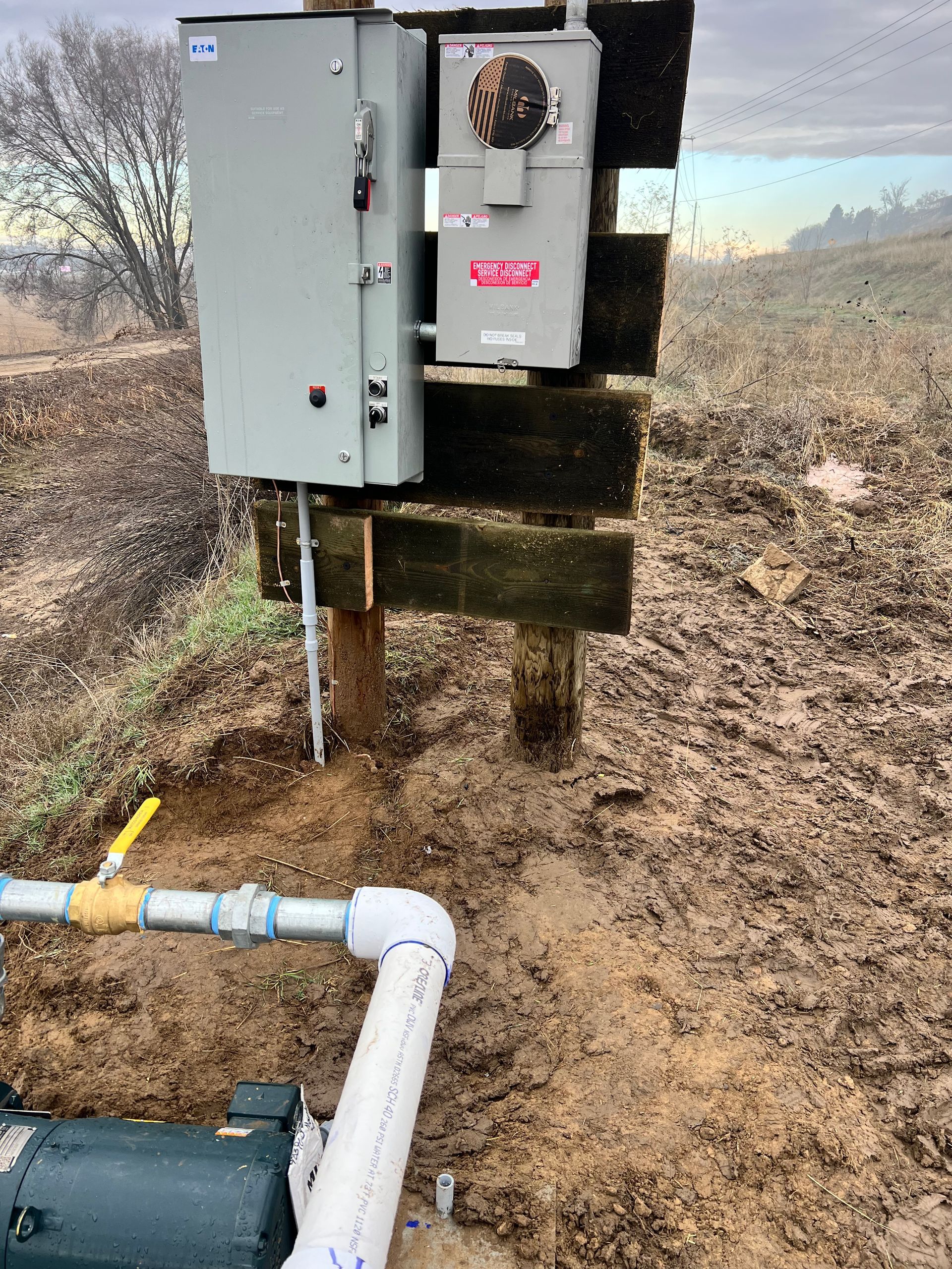 Utility meter and electrical boxes mounted on a wooden post beside a pipe in a dry field