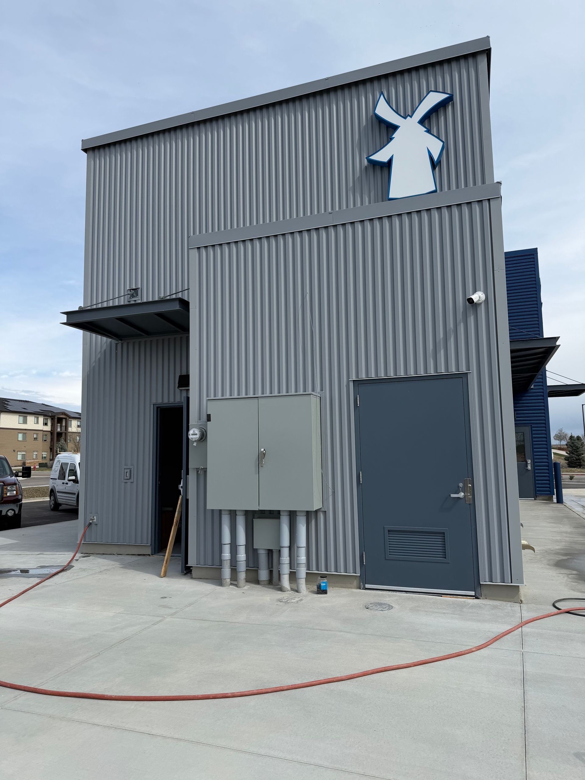 Gray corrugated metal building with a windmill logo, utility panels, and a blue door in an industrial lot