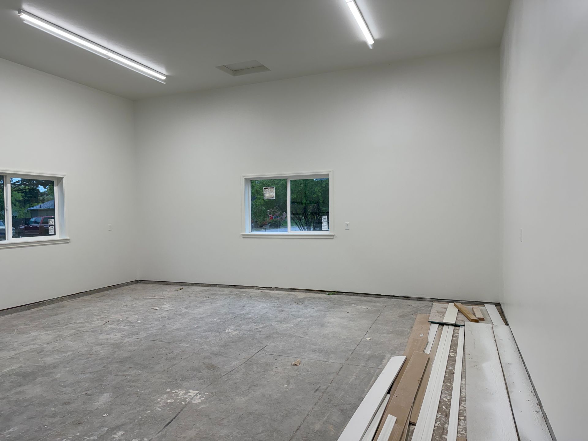 Empty room with white walls, fluorescent lights, and two windows; construction materials lie on the floor.