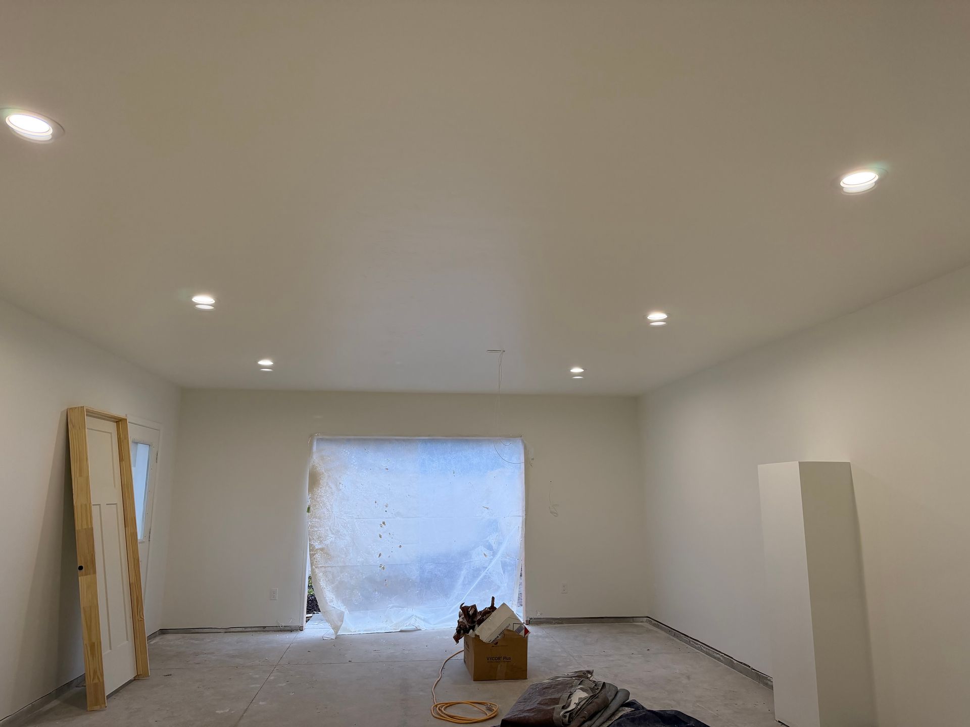 Empty white room under renovation with recessed ceiling lights and plastic-covered doorway at the far end