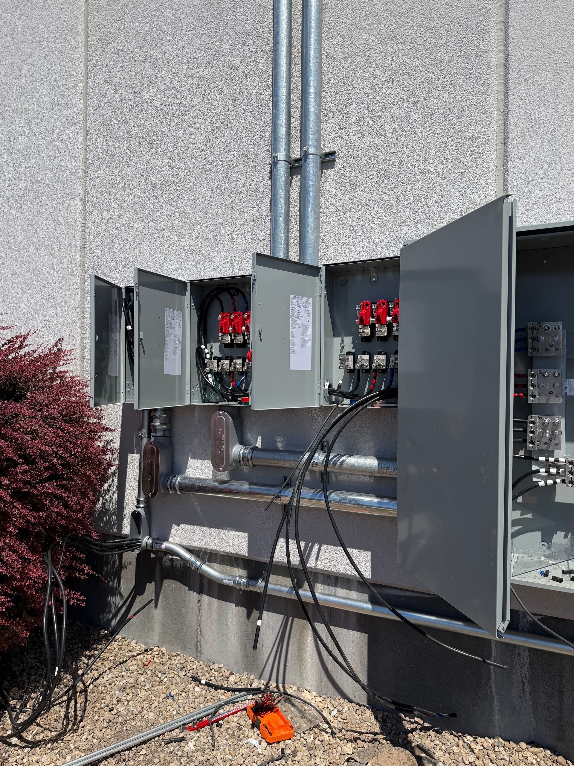 Open electrical panels and conduit mounted on an exterior wall with cables hanging below