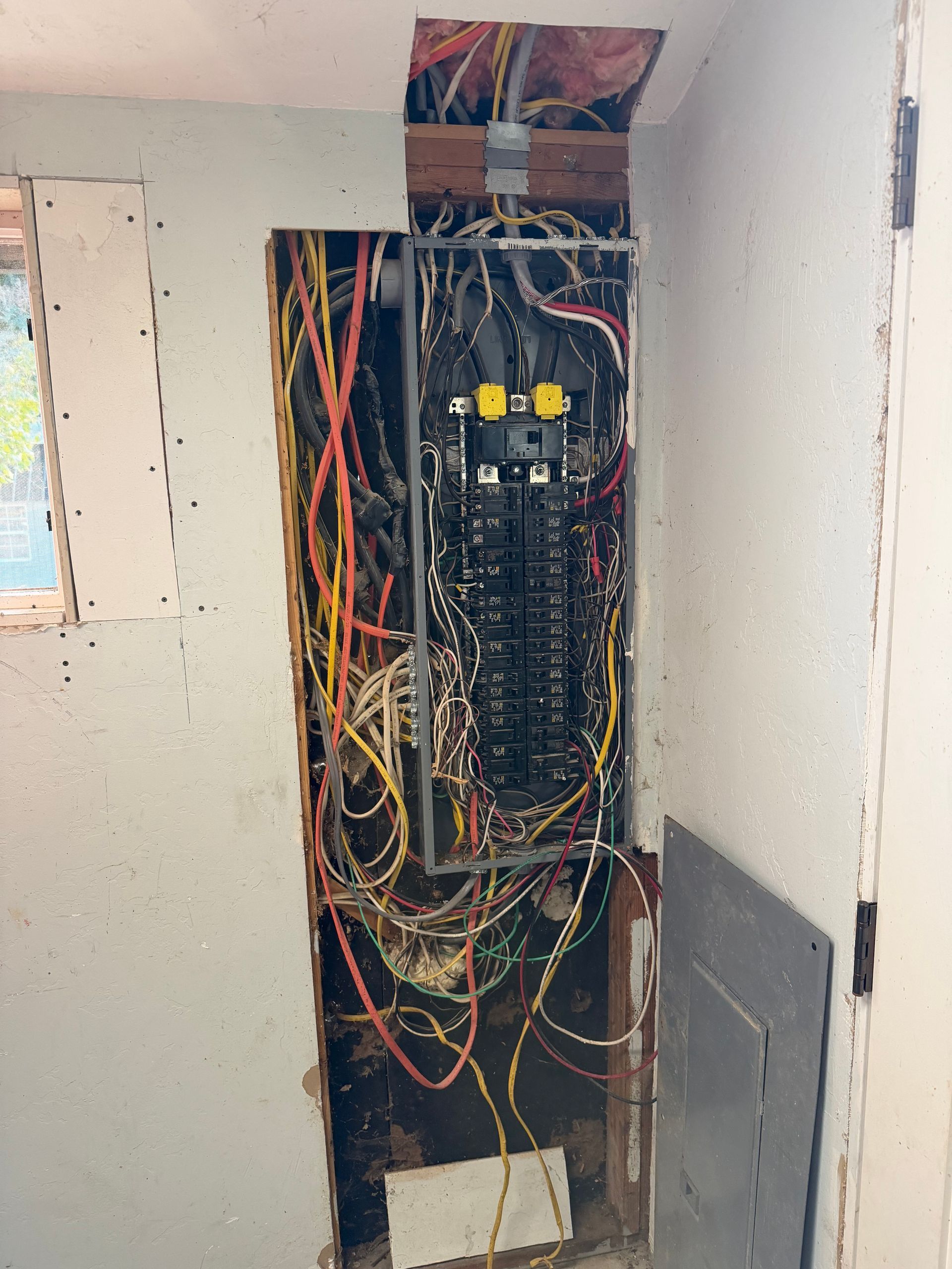 Exposed electrical breaker panel with tangled colored wires in a wall opening