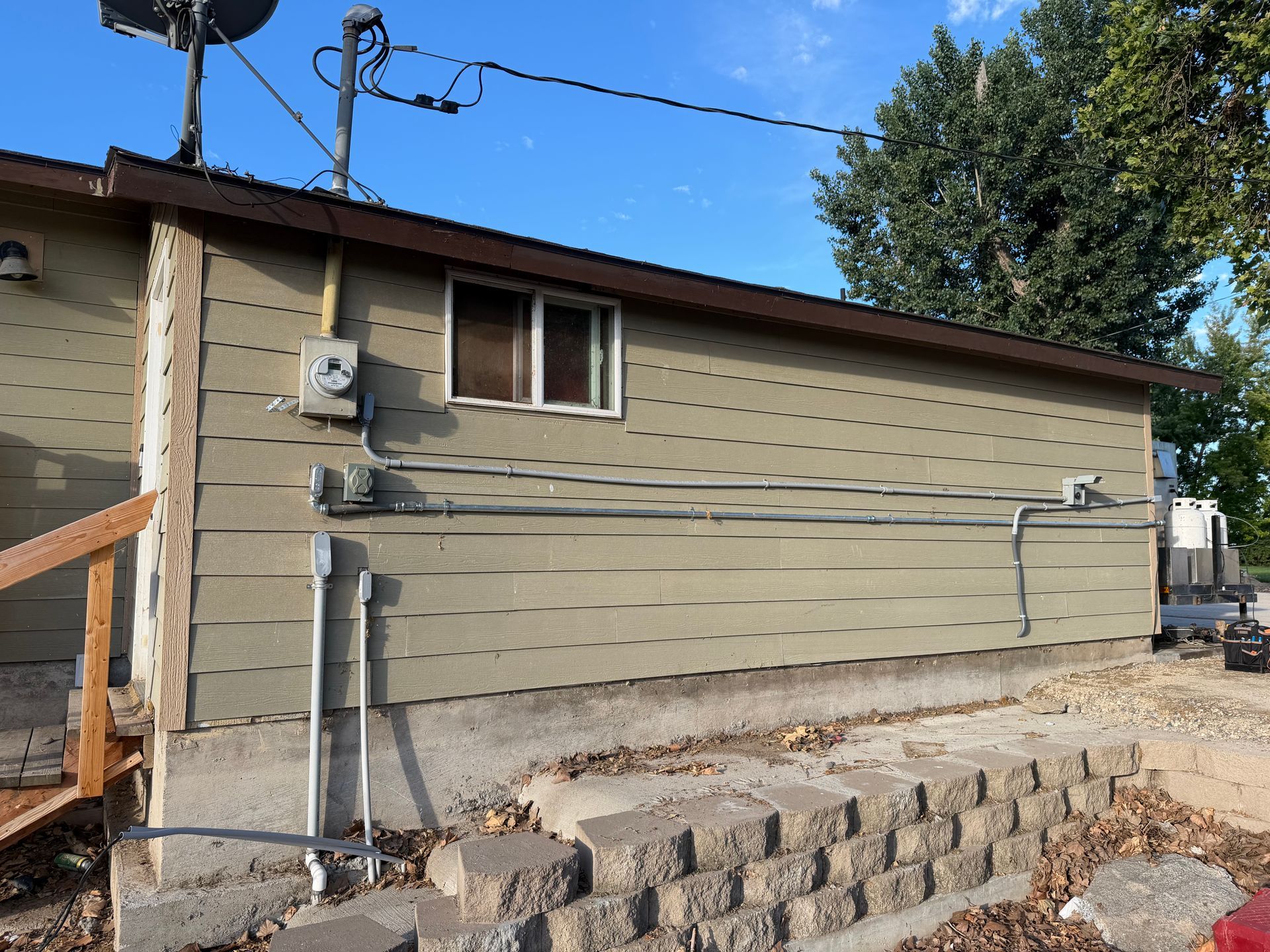 Side view of a beige house with utility meters, pipes, and a brick retaining wall under a blue sky