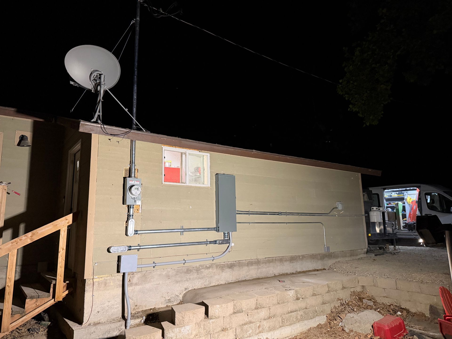 Nighttime exterior of a beige building with a satellite dish, utility boxes, and a parked vehicle beside steps