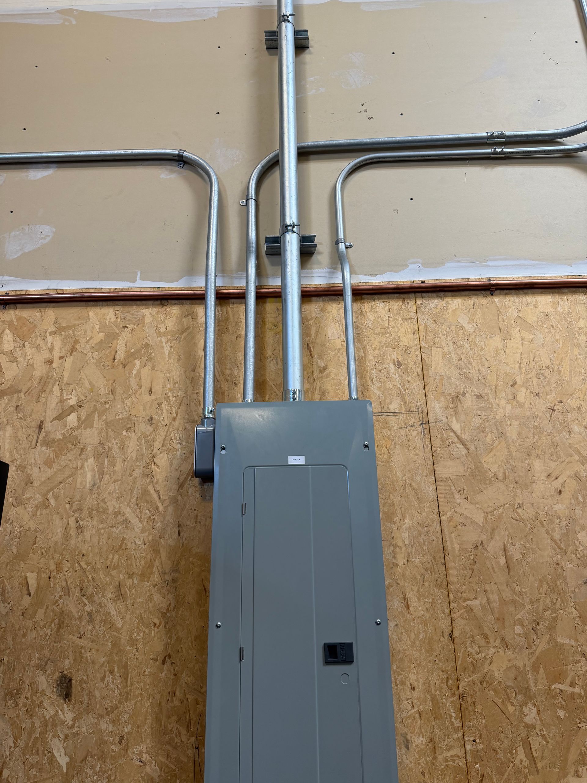Gray electrical breaker box with metal conduit pipes mounted on a wall of drywall and plywood.