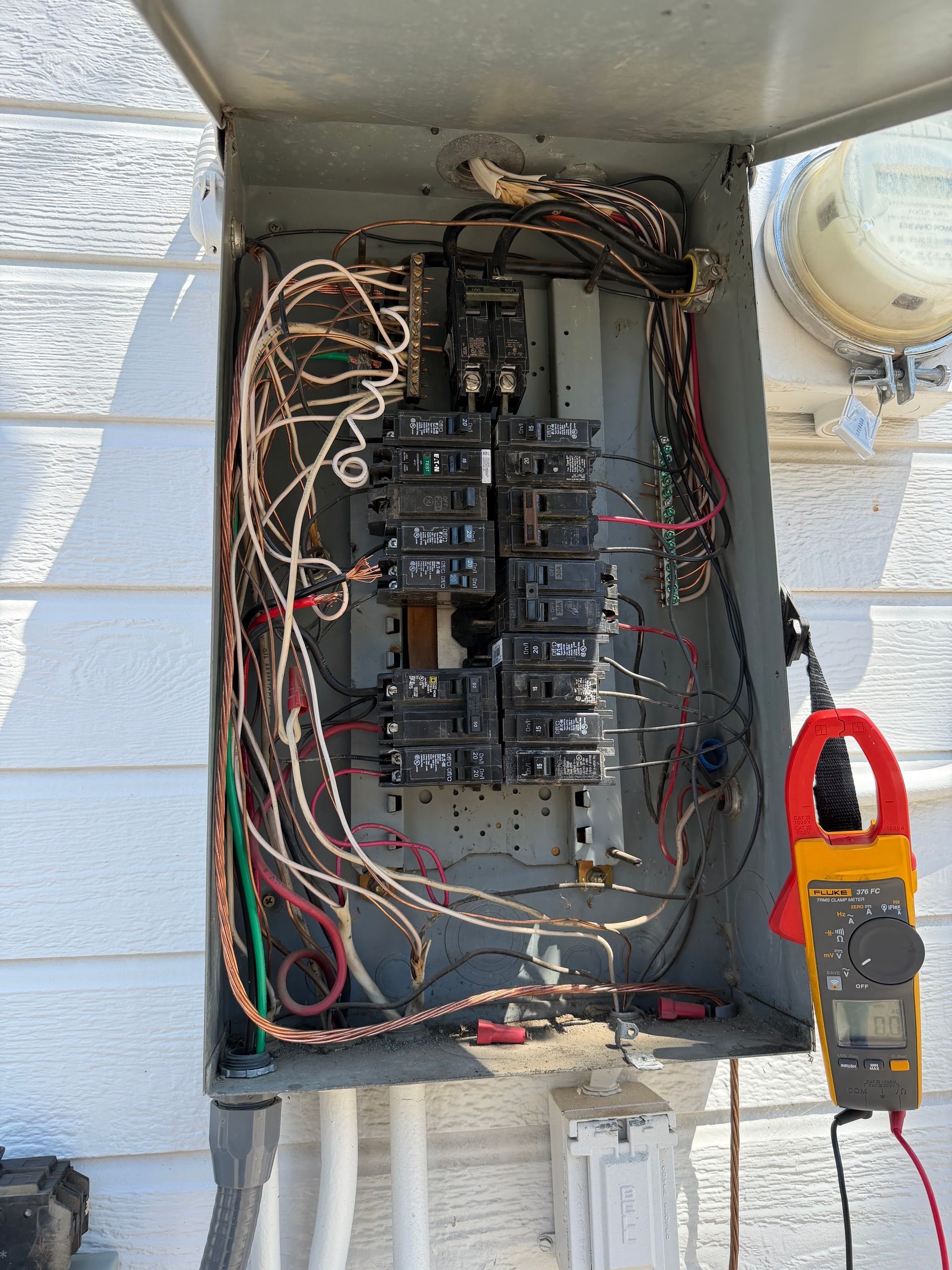 Open electrical breaker panel with exposed wiring and a clamp meter hanging on the right.