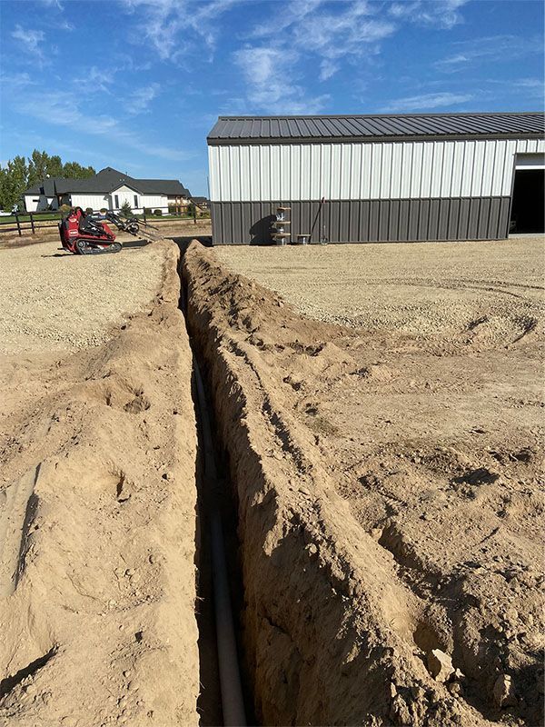 Deep trench dug beside a metal building on a gravel lot, with a small red machine in the distance.