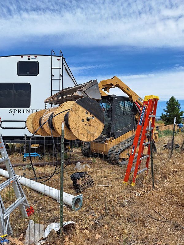 Yellow excavator lifting a large wooden spool beside a white RV in a dry construction area