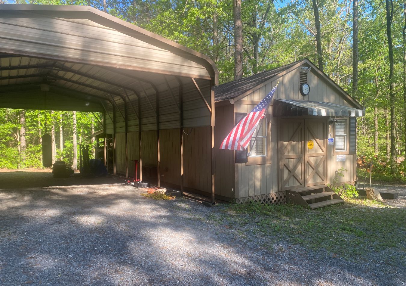 Gravel driveway beside a metal carport and small shed with an American flag, surrounded by trees.
