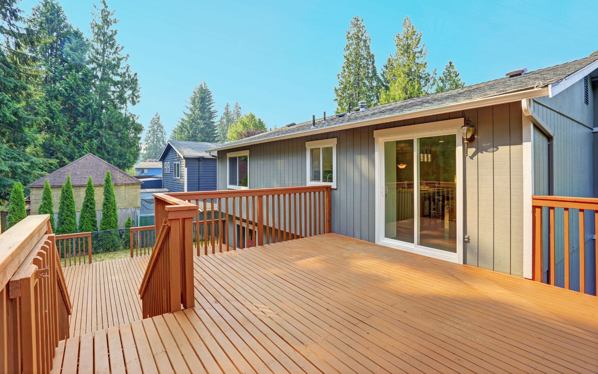 Wooden deck patio beside a house, with sliding glass doors and trees in the background.