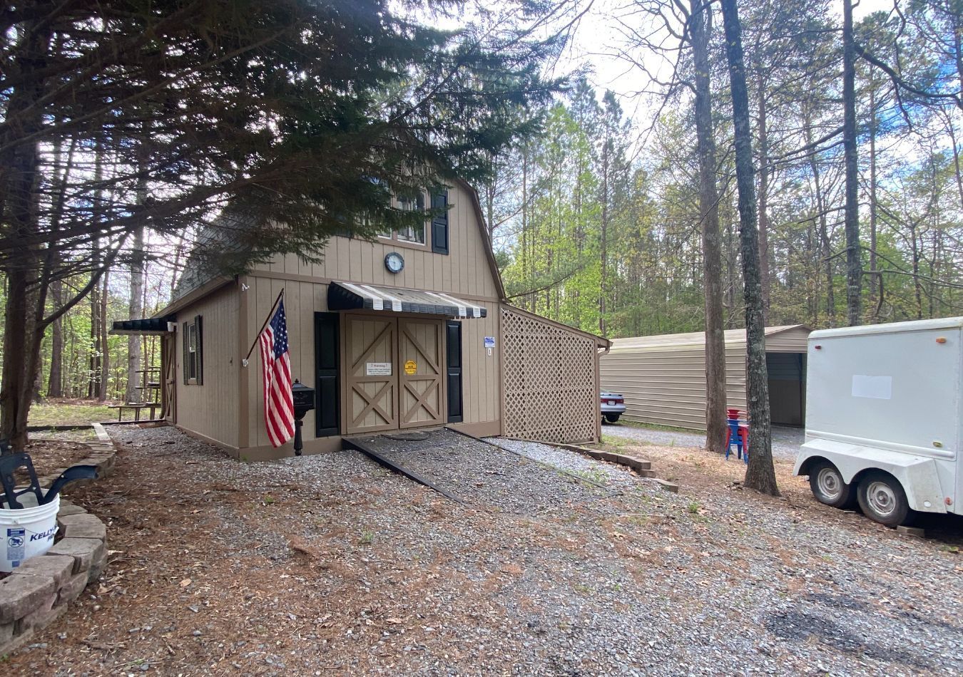 Small tan cabin in woods with American flag and parked white trailer in front