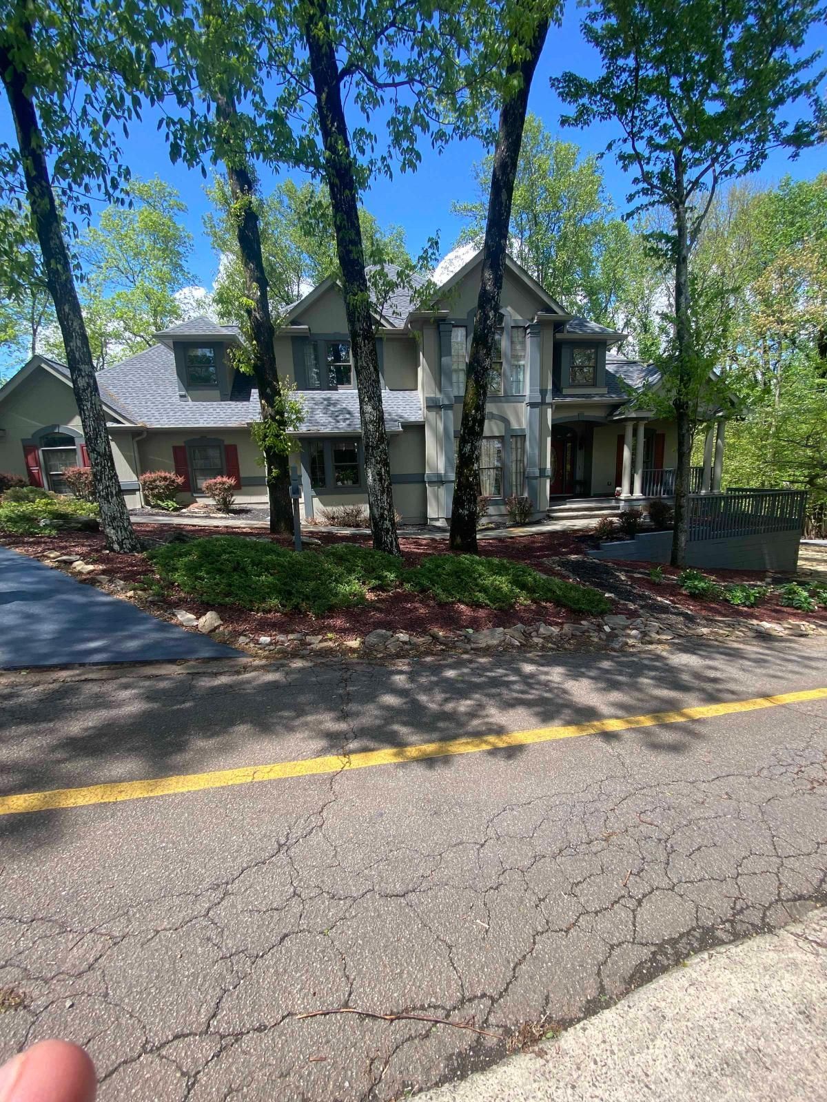 Two-story house with white siding and green trim beside a cracked road, shaded by tall trees.