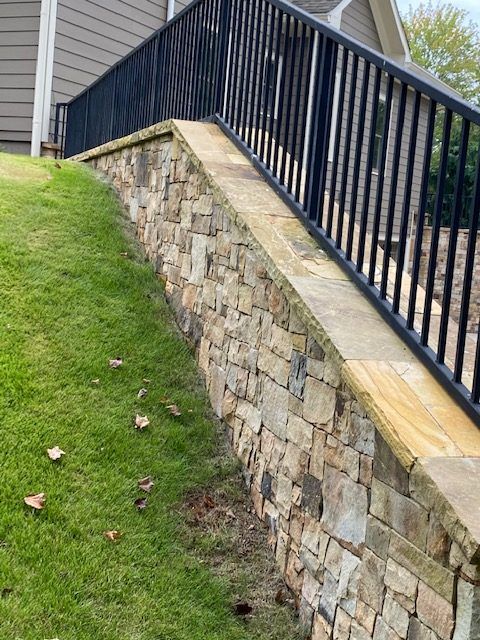 Stone retaining wall with black railing beside a sloped grassy yard and house siding