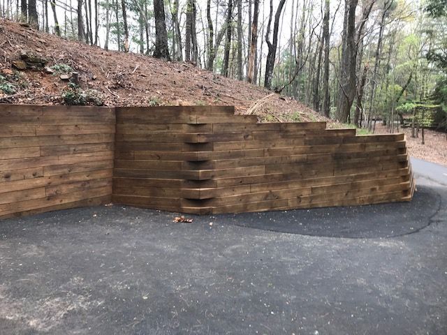 Wooden retaining wall with stone steps built into a wooded hillside beside an asphalt driveway.