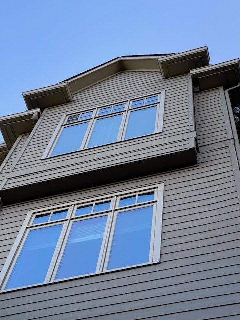 Upward view of a gray house exterior with two white-trimmed windows against a blue sky