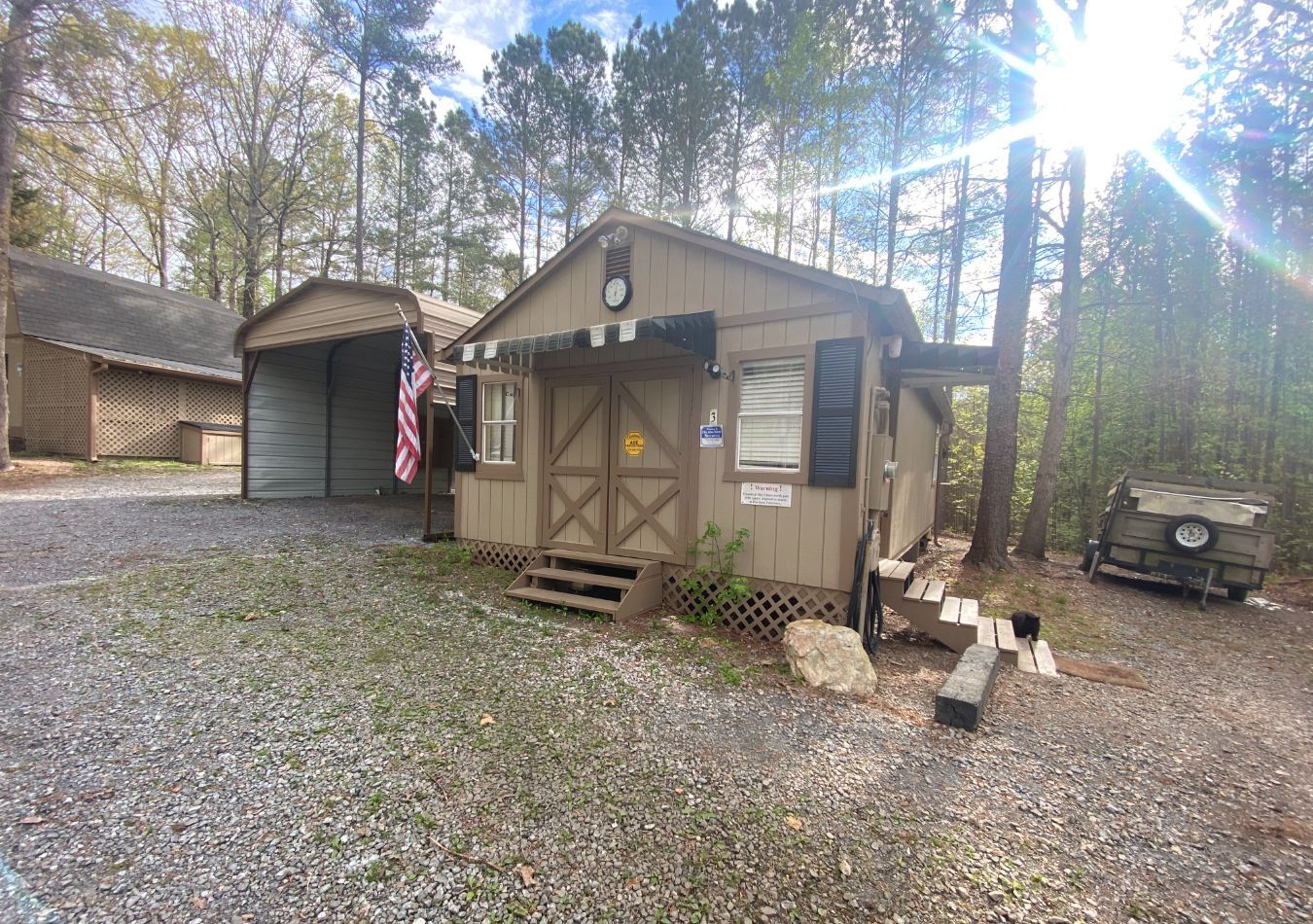 Small tan cabin with porch and American flag in a wooded gravel yard