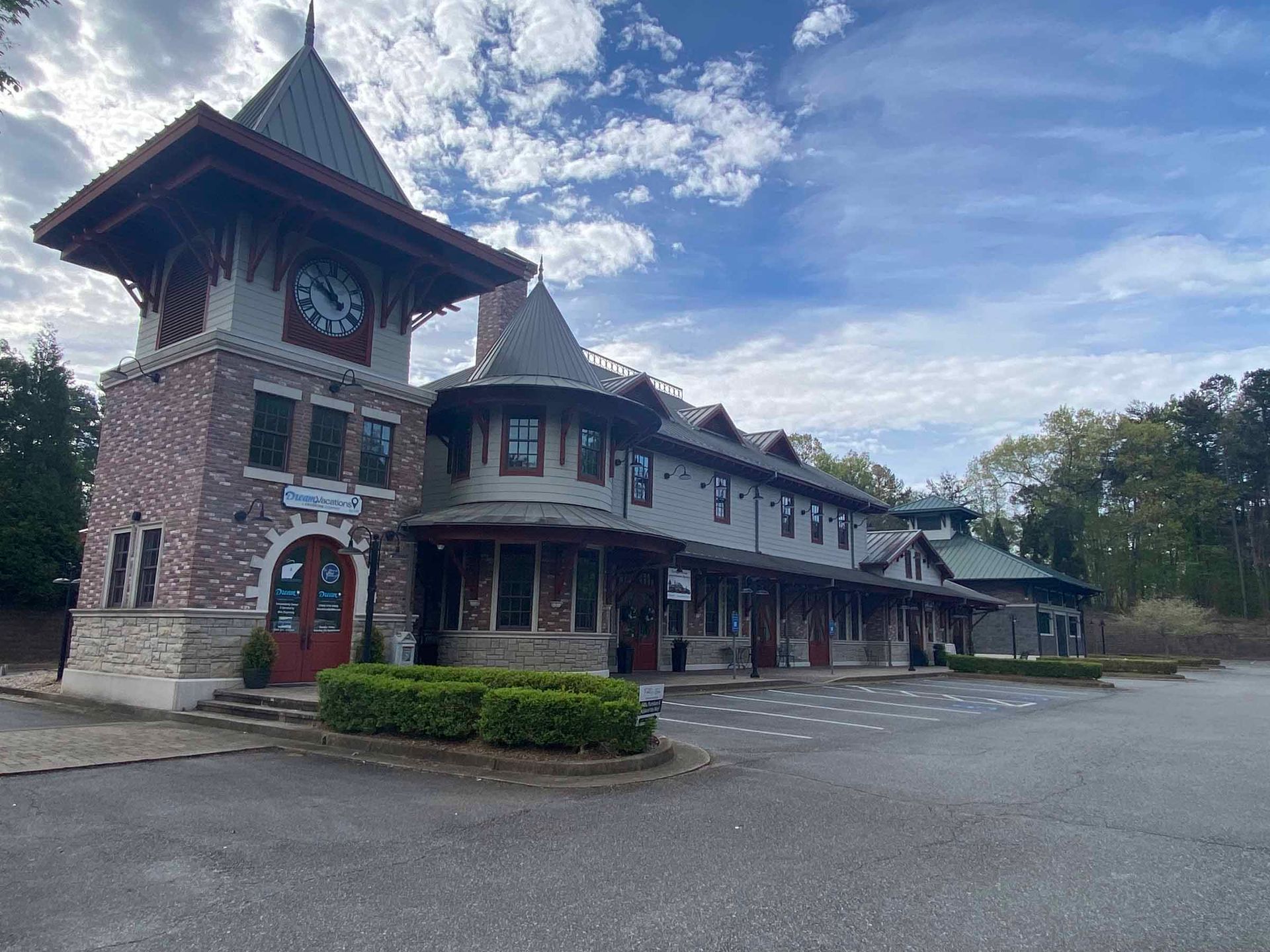 Stone building with a clock tower, covered porch, and empty parking lot under a cloudy blue sky