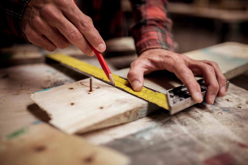 Hands measuring a wooden plank with a yellow tape measure and red pencil on a workbench.