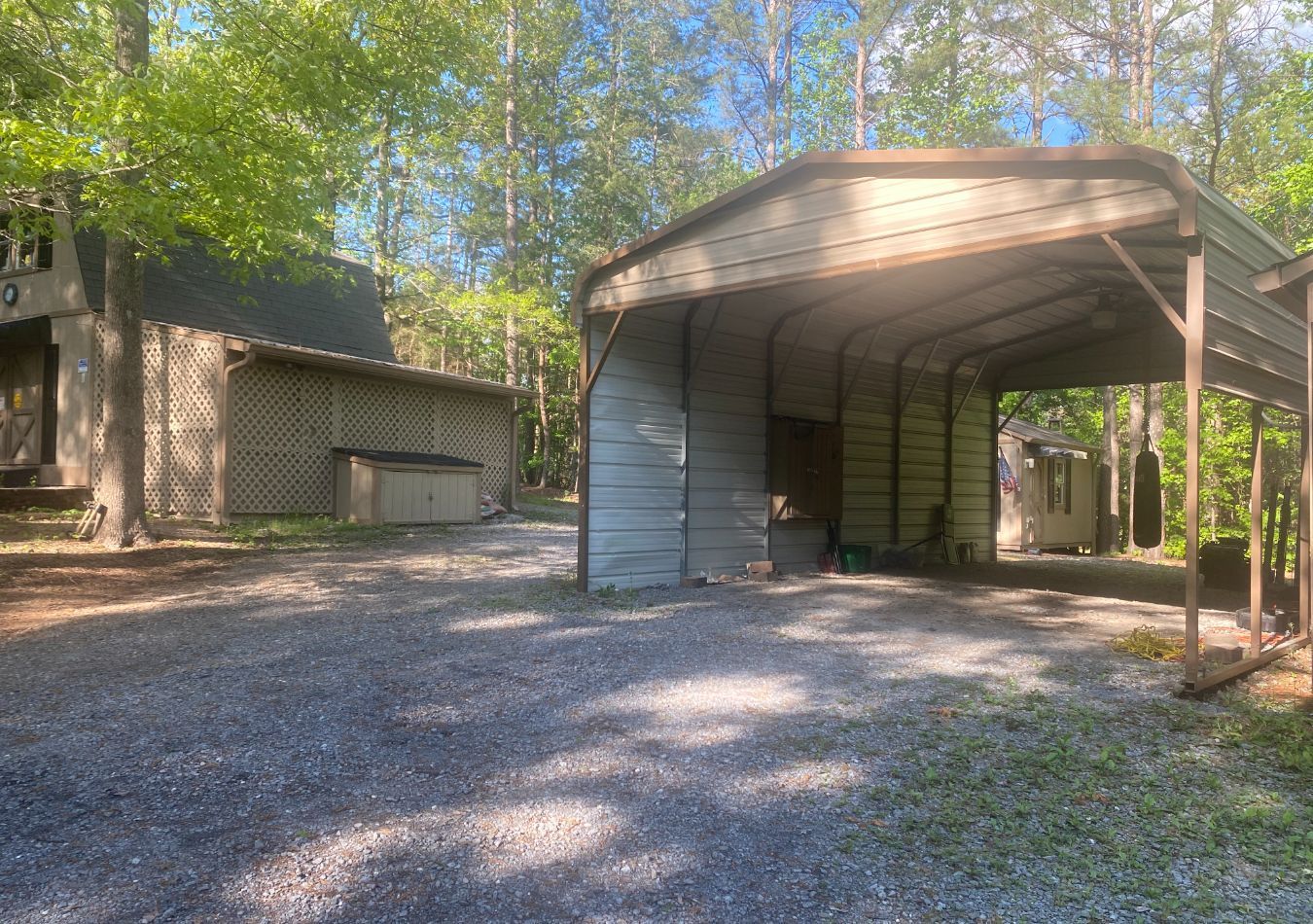 Gravel driveway with two metal carports and a small shed among trees in a wooded yard