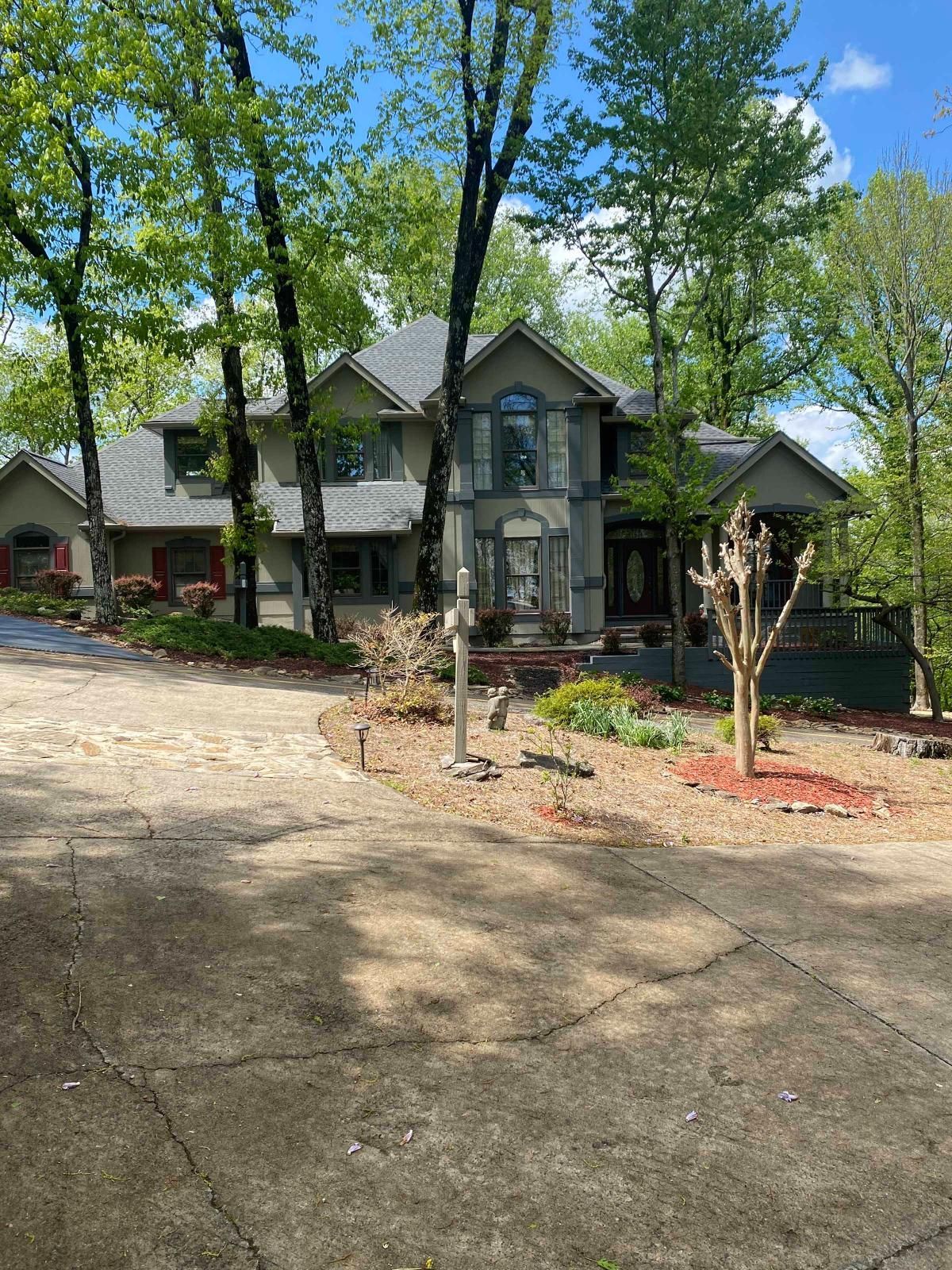 Two-story gray house with a front porch, trees, and a gravel driveway in sunlight