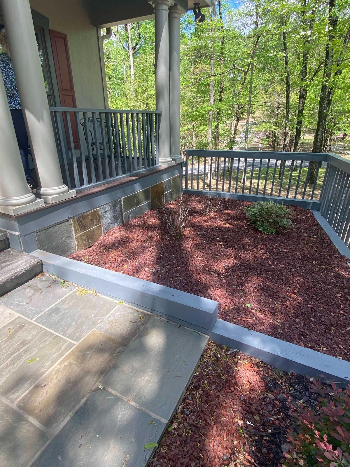 Front porch with gray stone patio, red mulch bed, blue-gray railing, and wooded yard beyond