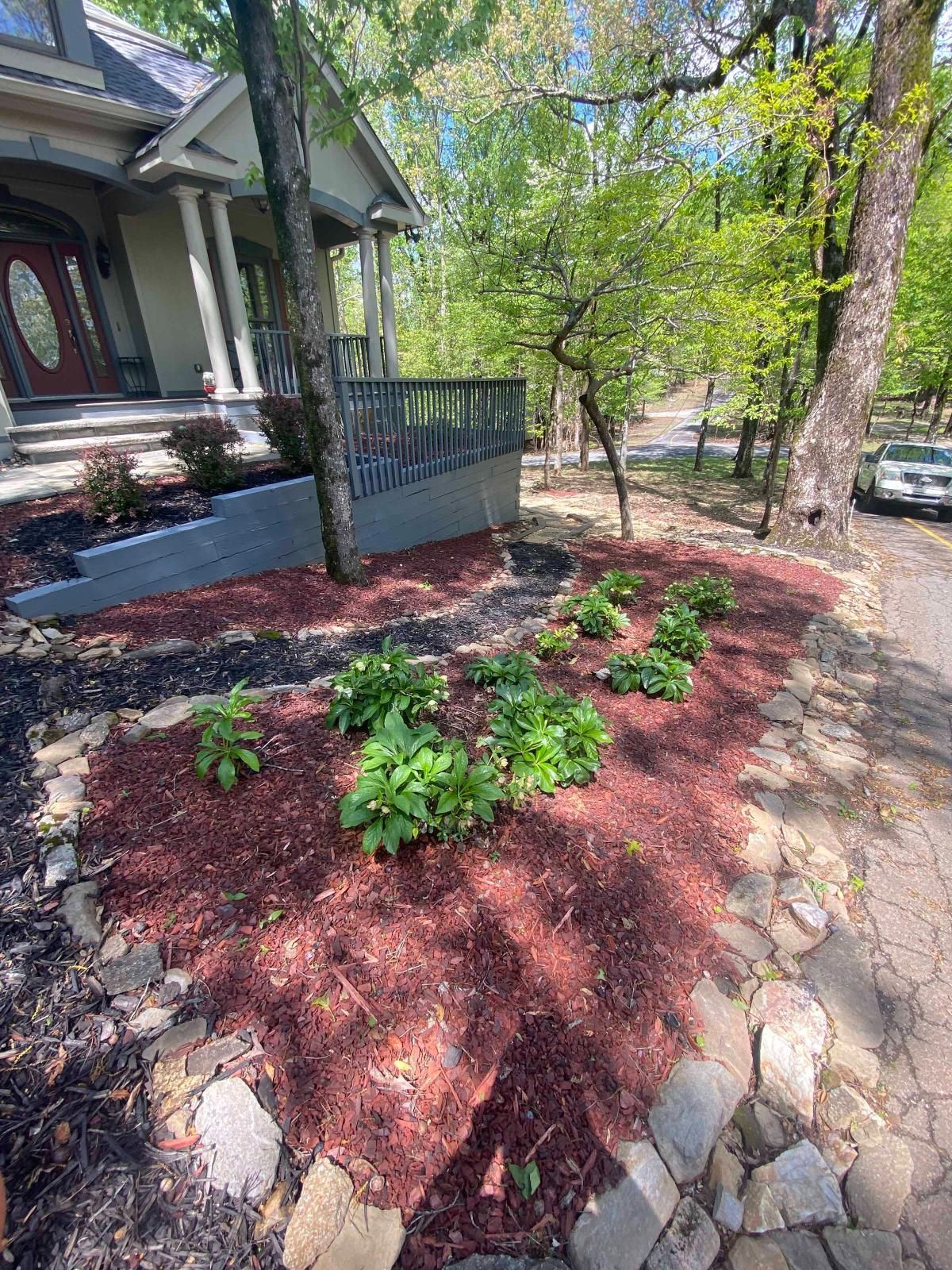 Front yard garden bed with green plants, red mulch, stone border, and a porch beside a tree-lined walkway